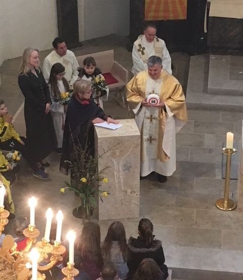 Ein Priester liest von einem Buch am Kirchenaltar. Menschen stehen und sitzen um ihn herum, darunter eine Frau mit Blumen.