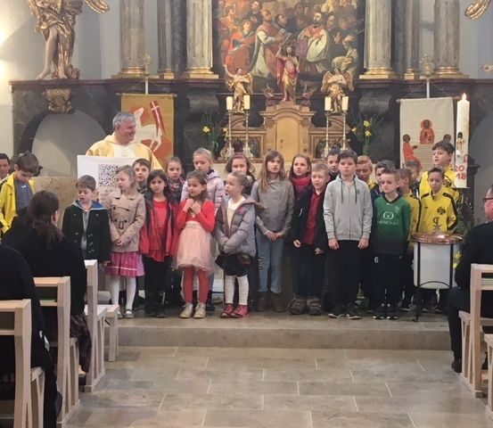Eine Gruppe von Kindern und Erwachsenen steht in einer Kirche, vor einem Altar mit Kerzen und einem Gemälde. Der Priester spricht zu ihnen.