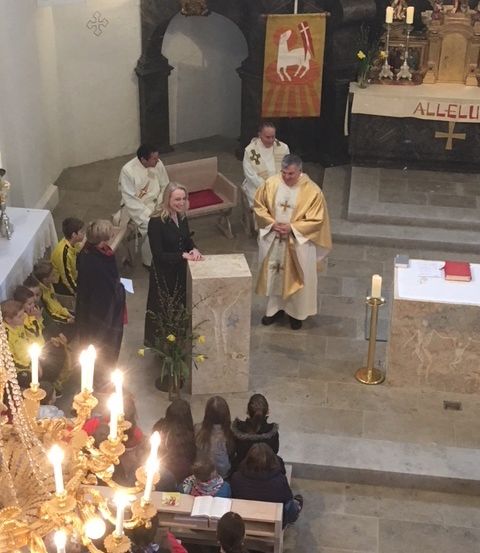 Ein Priester leitet einen Gottesdienst in einer Kirche. Die Menschen sitzen auf Kirchenbänken mit brennenden Kerzen. Eine Frau steht an einem Pult, und ein Banner hängt im Hintergrund.
