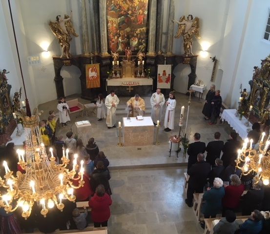 Eine religiöse Zeremonie findet in einer Kirche mit einem großen Altar, goldenen Kronleuchtern und Statuen statt. Priester und Messdiener sind am Altar, und viele Menschen stehen und sitzen.