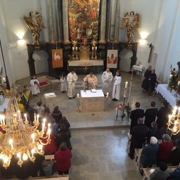 Eine religiöse Zeremonie findet in einer Kirche mit einem großen Altar, goldenen Kronleuchtern und Statuen statt. Priester und Messdiener sind am Altar, und viele Menschen stehen und sitzen.