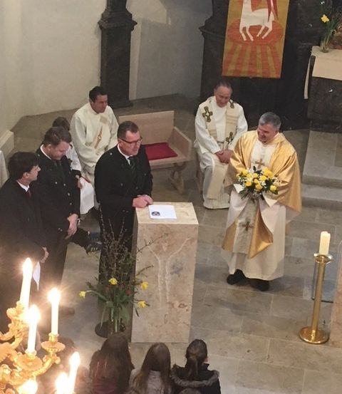 Ein Priester in einer goldenen Robe hält einen Blumenstrauß, während zwei Männer in Anzügen an einem Podest in einer Kirche stehen.