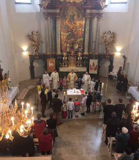 Eine religiöse Zeremonie in einer Kirche. Mehrere Personen stehen um einen Altar herum. Kerzen sind auf beiden Seiten angezündet.