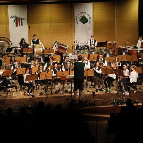 Ein großes Ensemble junger Musiker tritt auf der Bühne auf, mit Fahnen und Instrumenten im Hintergrund.