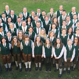Eine Gruppe von Menschen in traditioneller bayerischer Kleidung, die zusammen auf einem grünen Feld stehen und für ein Foto posieren.