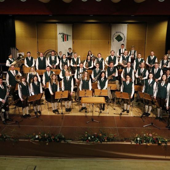 Ein großes Orchester in traditioneller grüner und weißer Kleidung posiert für ein Foto auf der Bühne, mit Musikinstrumenten und Bannern im Hintergrund.