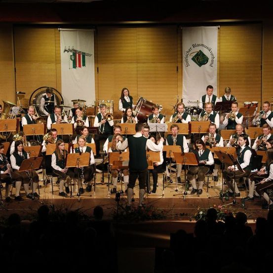 Ein Blasorchester spielt auf der Bühne mit verschiedenen Instrumenten, darunter Trompeten und Tuben, unter Bannern. Das Publikum sitzt vor der Bühne und beobachtet die Aufführung.