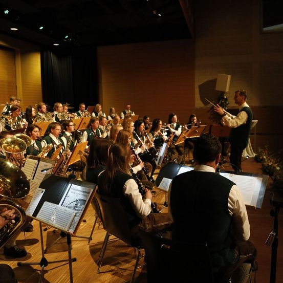 Ein großes Orchester spielt auf einer Bühne mit verschiedenen Instrumenten, darunter Trompeten, Posaunen und Flöten. Ein Dirigent leitet die Musiker. Einige Musiker sitzen, andere stehen.