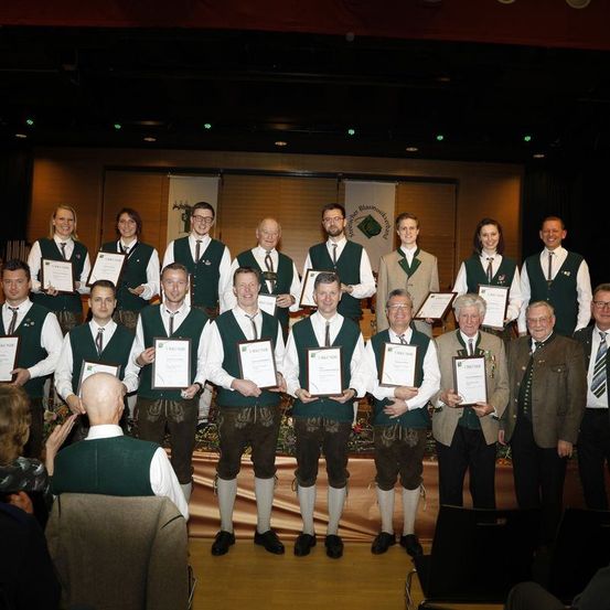 Eine Gruppe von Menschen, Männern und Frauen, steht zusammen auf einer Bühne. Sie tragen alle grüne Westen, Krawatten und Brillen. Sie halten Urkunden und posieren für ein Foto. Hinter ihnen stehen Stühle, ein Tisch und ein Vorhang. Es hängen auch Banner an der Wand.
