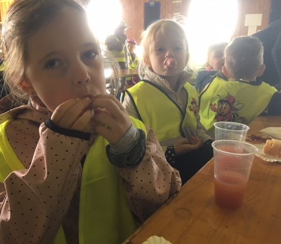 Kinder in gelben Westen sitzen an einem Tisch, essen und trinken. Ein Mädchen bläst eine Seifenblase. Andere Kinder und Erwachsene sind anwesend.