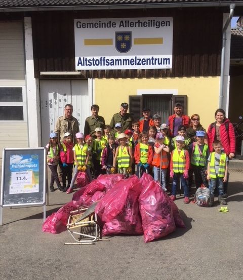 Eine Gruppe von Menschen, darunter Kinder, Erwachsene und einige uniformierte Personen, steht vor einem Gebäude mit der Aufschrift 'Altstoffsammlungszentrum' und posiert für ein Foto. Sie tragen Warnwesten und halten Müllsäcke.