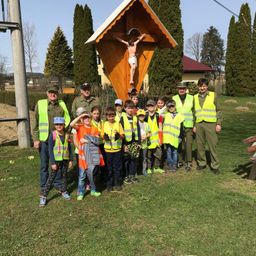 Eine Gruppe von Menschen, einschließlich Kindern, steht vor einem hölzernen Kruzifix in einem Park. Sie tragen alle neonfarbene Westen.