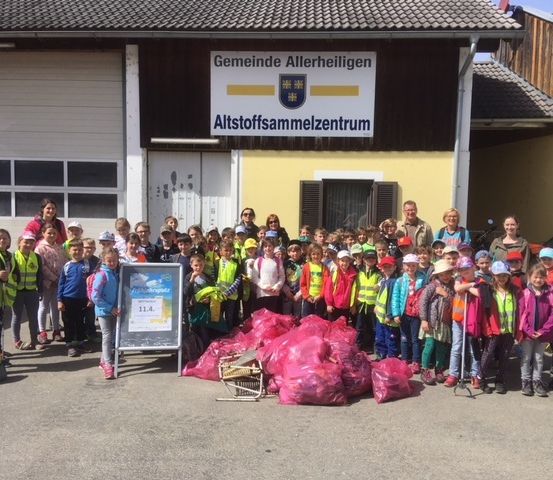 Eine Gruppe von Kindern in Sicherheitswesten steht vor einem Gebäude mit einem Schild, auf dem 'Gemeinde Allerheiligen Abfallstoffsammlungszentrum' steht. Sie posieren für ein Foto.
