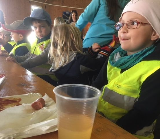 Mehrere Kinder in reflektierenden Westen sitzen an einem Tisch und essen. Ein Junge mit Brille trägt eine Mütze.