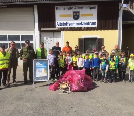 Eine Gruppe von Erwachsenen und Kindern in Warnwesten posiert vor einem Gebäude mit der Aufschrift 'Altstoffsammelzentrum' und einem großen Haufen Plastiktüten auf dem Boden.