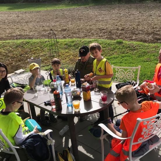 Kinder in gelben Westen sitzen um einen Tisch mit Getränken und Essen, wahrscheinlich auf einem Bauernhof. Ein Erwachsener in Uniform und Hut steht hinter ihnen.