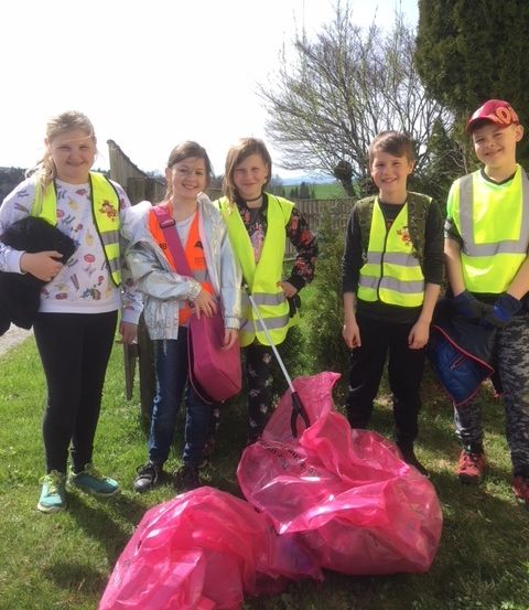 Fünf Kinder in Reflektorwesten stehen auf einem Grasfeld und halten Plastiktüten in den Händen. Sie scheinen an einer Aufräumaktion teilzunehmen. Im Hintergrund sind Bäume und ein Zaun zu sehen.