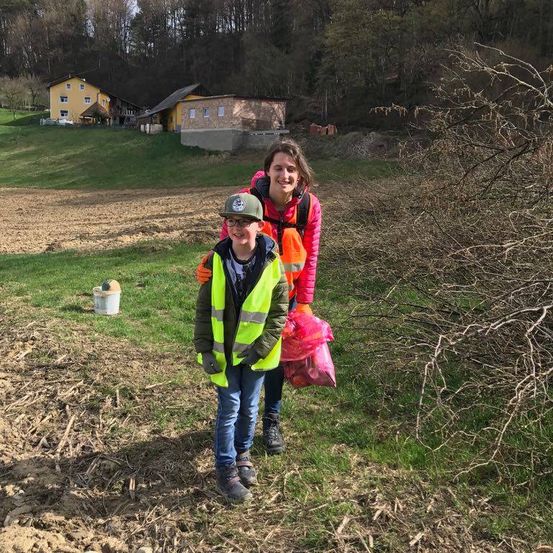 Eine Frau und ein Junge stehen in einem Feld und lächeln für ein Foto. Der Junge trägt eine reflektierende Weste. Dahinter steht ein Eimer und ein Haus auf einem Hügel.