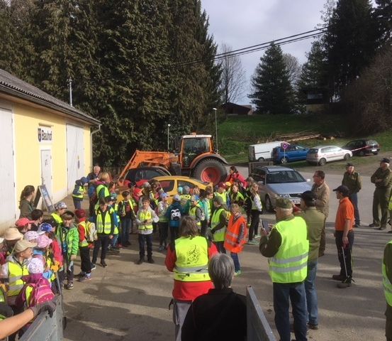 Eine Gruppe von Kindern und Erwachsenen in Warnwesten steht vor einem Gebäude, in der Nähe geparkter Fahrzeuge und eines Maschinenfahrzeugs.