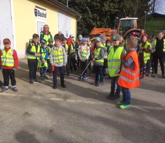 Eine Gruppe von Kindern in Sicherheitswesten und Mützen steht vor einem Gebäude mit einem geparkten Traktor und Auto in der Nähe.