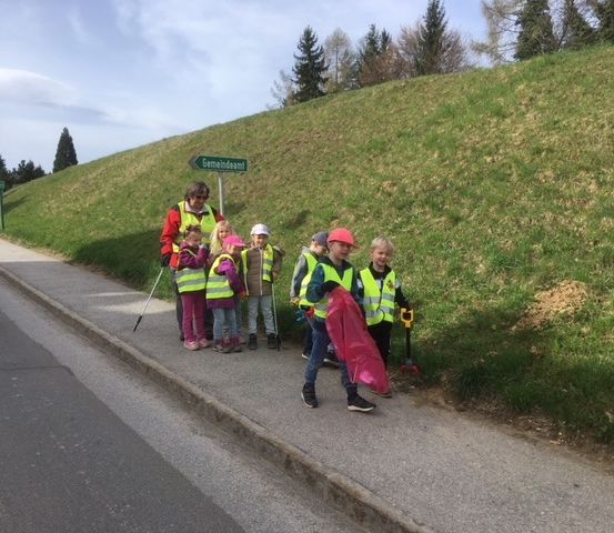 Eine Gruppe von Kindern und einem Erwachsenen in Warnwesten steht am Straßenrand und hält einen Plastikbeutel und ein Werkzeug.
