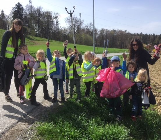 Eine Gruppe von Kindern und zwei Erwachsenen in gelben Westen sammelt Müll entlang einer Straße mit grünem Gras und Bäumen im Hintergrund.