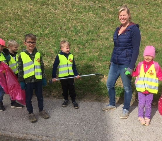 Eine Frau und fünf Kinder stehen auf einem Bürgersteig, tragen Sicherheitswesten und halten Müllsäcke. Sie scheinen an einer Gemeindereinigung teilzunehmen.
