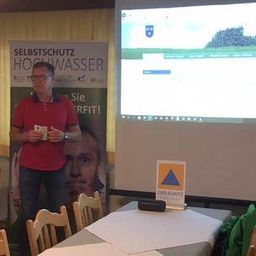 Ein Mann in einem roten Shirt steht vor einer Projektionsleinwand, die eine Landschaft mit einem Wasserzeichen zeigt. Ein großes Poster ist hinter ihm. Er hält eine Karte und schaut das Publikum an. Es gibt Tische und Stühle im Raum.