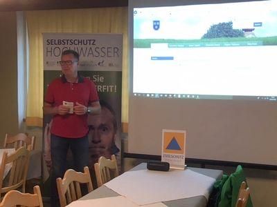 Ein Mann in einem roten Shirt steht vor einer Projektionsleinwand, die eine Landschaft mit einem Wasserzeichen zeigt. Ein großes Poster ist hinter ihm. Er hält eine Karte und schaut das Publikum an. Es gibt Tische und Stühle im Raum.