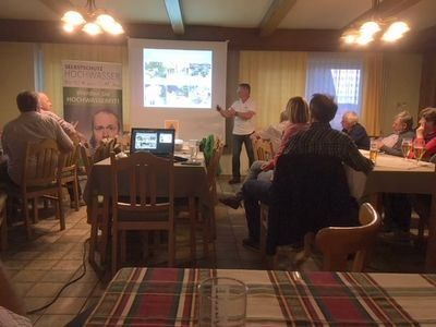 Eine Gruppe von Menschen sitzt um Tische herum in einem Raum und hört einem Mann zu, der vor einer Projektionsleinwand steht. Die Leinwand zeigt ein Bild. Der Raum hat Deckenleuchten und ein Fenster.
