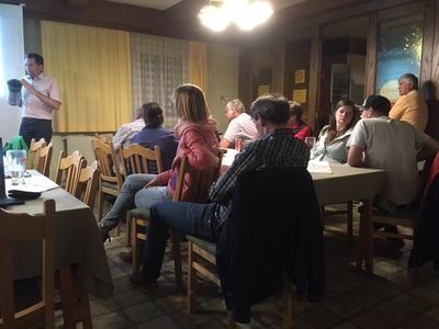 Eine Gruppe von Menschen sitzt um Tische herum in einem Raum. Ein Mann steht vorne und hält möglicherweise eine Präsentation. Der Raum hat Holzstühle, Tische und eine Projektionsleinwand. Die Wände sind mit Gemälden und Vorhängen dekoriert.