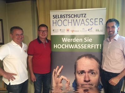 Vier Männer stehen neben einem Plakat, auf dem das Gesicht eines Mannes unter Wasser abgebildet ist, mit dem Text 'Werden Sie Hochwasserfit!' in prominenter Darstellung. Das Plakat hat einen grünen Hintergrund und weißen Text.