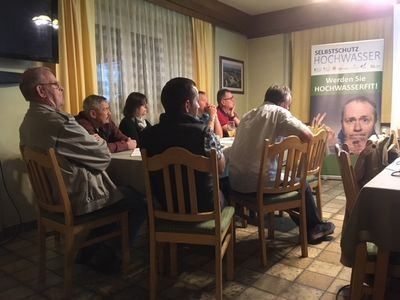 Eine Gruppe von Menschen sitzt um einen Tisch in einem Raum. Ein Banner mit einem Männergesicht und Text ist an der Wand. Der Raum hat Holzstühle, einen Tisch und einen gefliesten Boden. Ein Fernseher ist an der Wand montiert.
