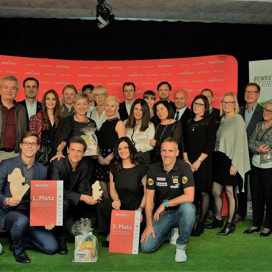 Eine Gruppe von Menschen posiert für ein Foto bei einer Veranstaltung. Einige halten Trophäen. Ein Banner ist hinter ihnen platziert, und einige Werbetafeln sind an der Wand angebracht.