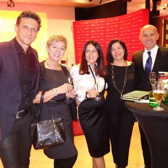 Fünf Personen posieren für ein Foto, Champagnergläser in der Hand, lächelnd. Ein Tisch mit einem grünen Getränk und einer Schachtel mit Karomuster steht dahinter.