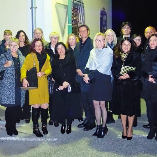 Eine Gruppe von Menschen, sowohl Männer als auch Frauen, posiert für ein Foto draußen bei Nacht. Sie sind in Winterkleidung gekleidet, einige mit Brillen. Eine Frau hält ein kariertes Buch.