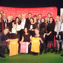 Eine Gruppe von Menschen steht vor einem roten Hintergrund und hält orangefarbene Hemden, möglicherweise zur Feier eines Ereignisses. Einige tragen Brillen und Schmuck, und zwei Personen halten Auszeichnungen. Hinter ihnen ist ein roter Banner mit Logos und Text zu sehen.