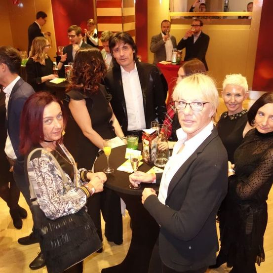 Eine Gruppe von Menschen versammelt sich in einem Raum, einige stehen und einige sitzen. Eine Frau hält ein Weinglas, während eine andere ein Glas Orangensaft hält. Ein Mann in der Mitte schaut auf etwas auf dem Tisch. Hinter ihnen stehen Menschen, die etwas trinken.