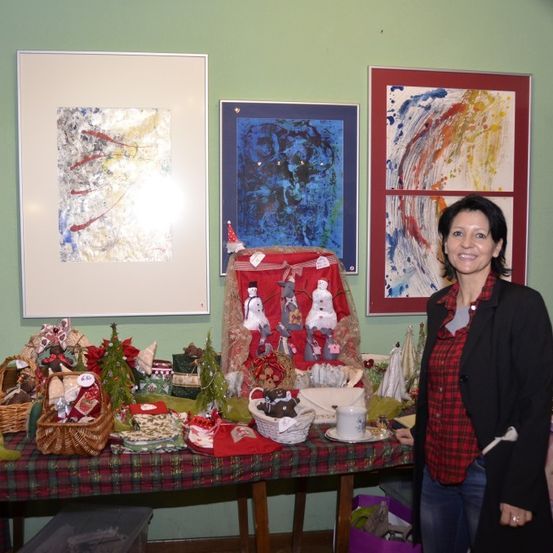 Eine Frau steht neben einem Tisch, der mit einem rot-schwarz karierten Tuch bedeckt ist und mit verschiedenen Weihnachtsdekorationen geschmückt ist. Hinter ihr hängen drei gerahmte Kunstwerke an einer grünen Wand.