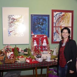 Eine Frau steht neben einem Tisch, der mit einem rot-schwarz karierten Tuch bedeckt ist und mit verschiedenen Weihnachtsdekorationen geschmückt ist. Hinter ihr hängen drei gerahmte Kunstwerke an einer grünen Wand.