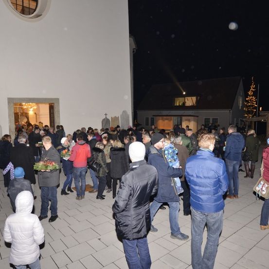 Viele Menschen haben sich bei Nacht vor einer Kirche versammelt, halten Blumen und tragen Winterkleidung. Ein Weihnachtsbaum ist im Hintergrund zu sehen.