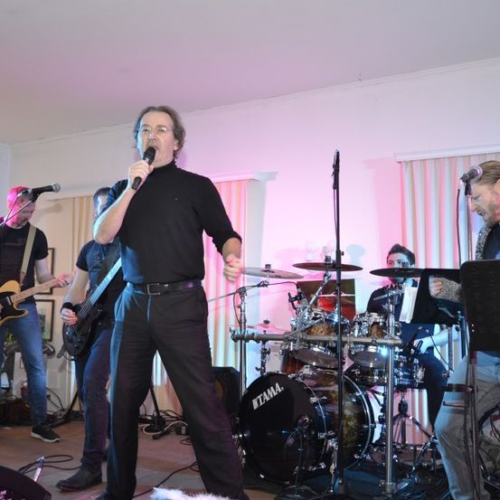 Eine Band spielt auf der Bühne. Der Leadsänger, der ein schwarzes Rollkragenshirt und eine Brille trägt, singt in ein Mikrofon. Hinter ihm steht ein Schlagzeuger, der ein Schlagzeug spielt, und zwei Gitarristen, die ihre Gitarren spielen. Vor ihnen befinden sich zwei Mikrofone auf Ständern. Die Bühne hat eine rosa Wand und Vorhänge.