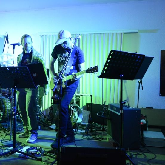 Zwei Männer stehen auf einer Bühne und spielen in einem schwach beleuchteten Raum. Einer spielt die Gitarre, während der andere das Schlagzeug spielt. Vor ihnen steht ein Mikrofon.