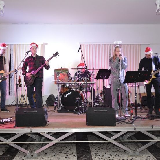 Eine Band spielt auf der Bühne mit Gitarren, Trommeln und einem Mikrofon. Die Mitglieder tragen Weihnachtsmützen. Die Bühne hat Lautsprecher und einen Hintergrund aus Vorhängen.
