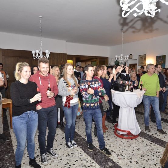 Eine Gruppe von Menschen steht in einem Raum, wahrscheinlich auf einer Party. Sie alle lächeln und einige halten Flaschen. Ein Tisch mit weißer Tischdecke und Flaschen steht vor ihnen.