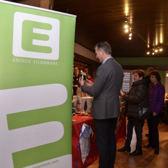 Ein Mann in einem Anzug betrachtet ein Telefon in der Nähe eines Banners für Energie Steiermark, mit mehreren Personen, die hinter ihm in einem Raum mit einem Tisch und dekorativen Gegenständen stehen.