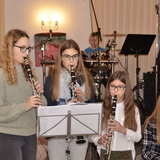 Drei Mädchen spielen Klarinetten in einem Raum mit Musikinstrumenten und einem Schlagzeug. Ein junger Junge spielt im Hintergrund das Schlagzeug.