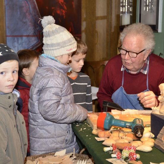 Ein älterer Mann schnitzert Holz an einem Tisch, während Kinder zusehen. Ein Kind trägt eine grau-weiße Mütze.
