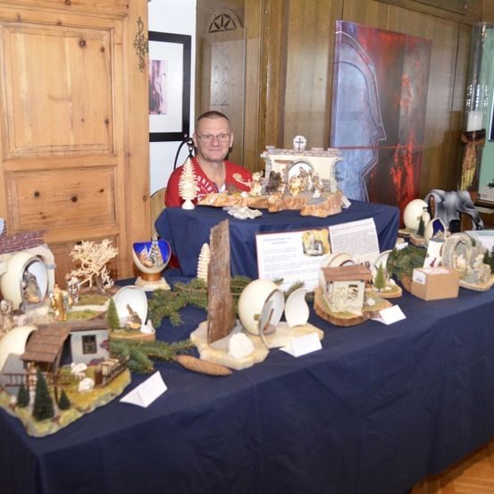 Ein Mann sitzt hinter einem mit einem blauen Tuch bedeckten Tisch und zeigt verschiedene Figuren und Kunstwerke. Der Tisch ist voll mit verschiedenen Gegenständen, darunter Skulpturen und ein Kreuz.