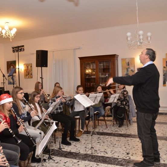 Ein Mann dirigiert eine Gruppe junger Musiker, die Flöten und Klarinetten spielen, vor dem Hintergrund eines Raums mit weißen Wänden, einer Kronleuchter und einem hölzernen Schrank.